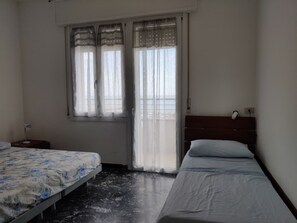 Ein Schlafzimmer mit zwei Betten, einem Fenster mit durchsichtigen Vorhängen, einem Nachttisch und einem Beistelltisch.