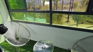 En balkong med två stolar och ett litet bord, fri utsikt över byggnader och palmer, samt konstgjord, grön gräsmatta.