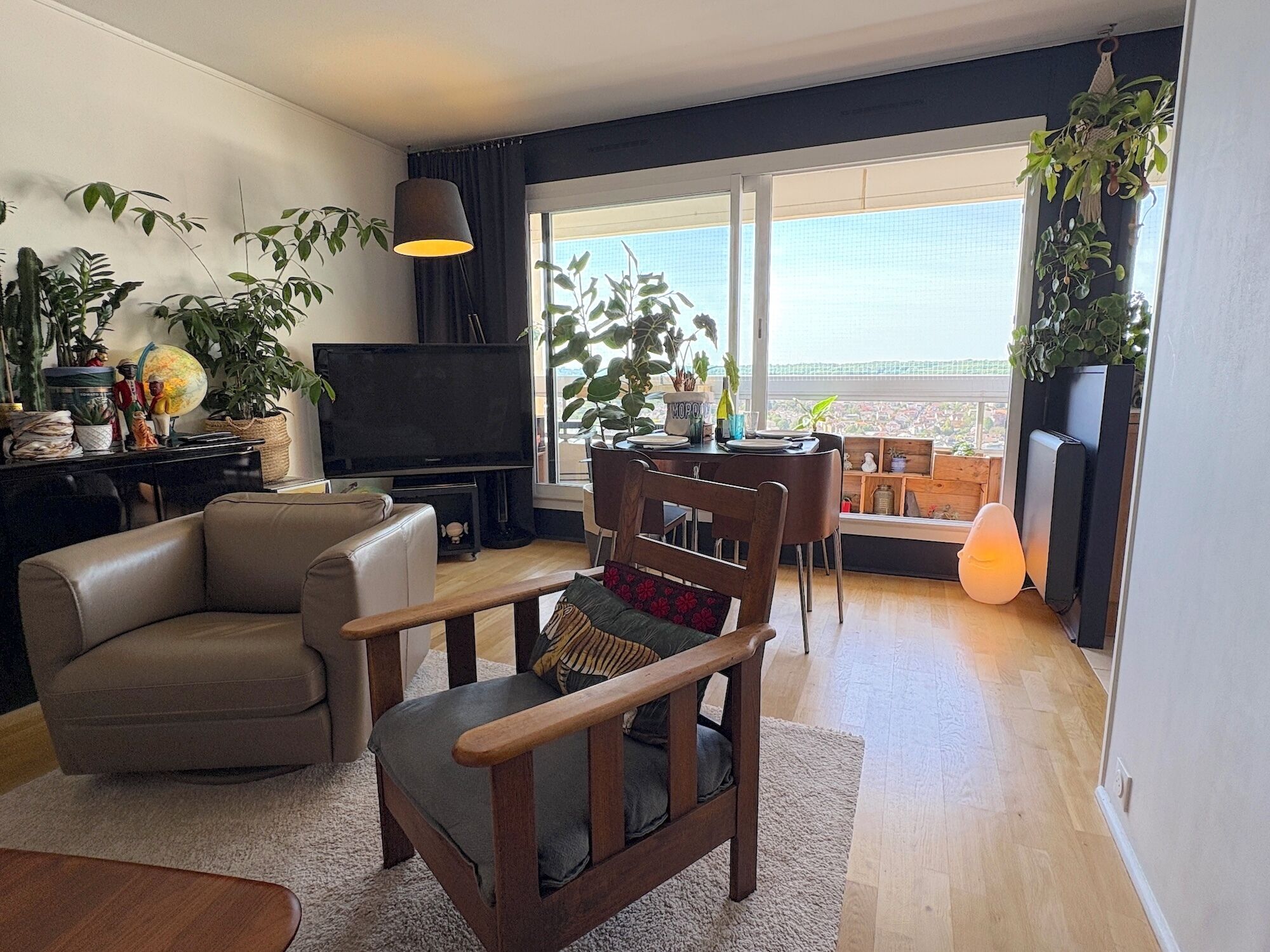 Un salon moderne comprenant un canapé, un fauteuil en bois, un téléviseur et un coin repas avec une table et des chaises. On y trouve également plusieurs plantes en pot et une grande fenêtre offrant une vue dégagée.