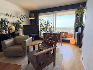 Un salon moderne comprenant un canapé, un fauteuil en bois, un téléviseur et un coin repas avec une table et des chaises. On y trouve également plusieurs plantes en pot et une grande fenêtre offrant une vue dégagée.