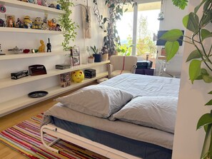 Une chambre à coucher avec un lit, des étagères remplies de divers objets, une chaise et une fenêtre ornée de plantes.