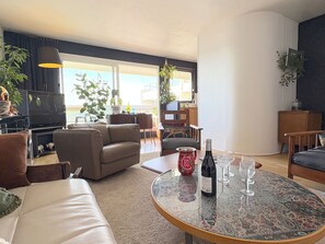 Un salon comprenant un canapé, des fauteuils, une table basse et un téléviseur. On y trouve également des plantes, un coin repas et une vue dégagée grâce à de grandes fenêtres.