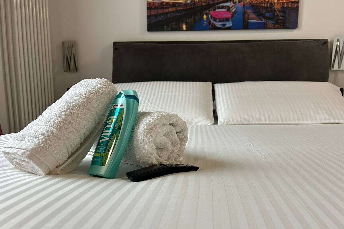 Un lit bien fait, avec deux oreillers, une serviette blanche roulée, une bouteille de shampoing et une télécommande.