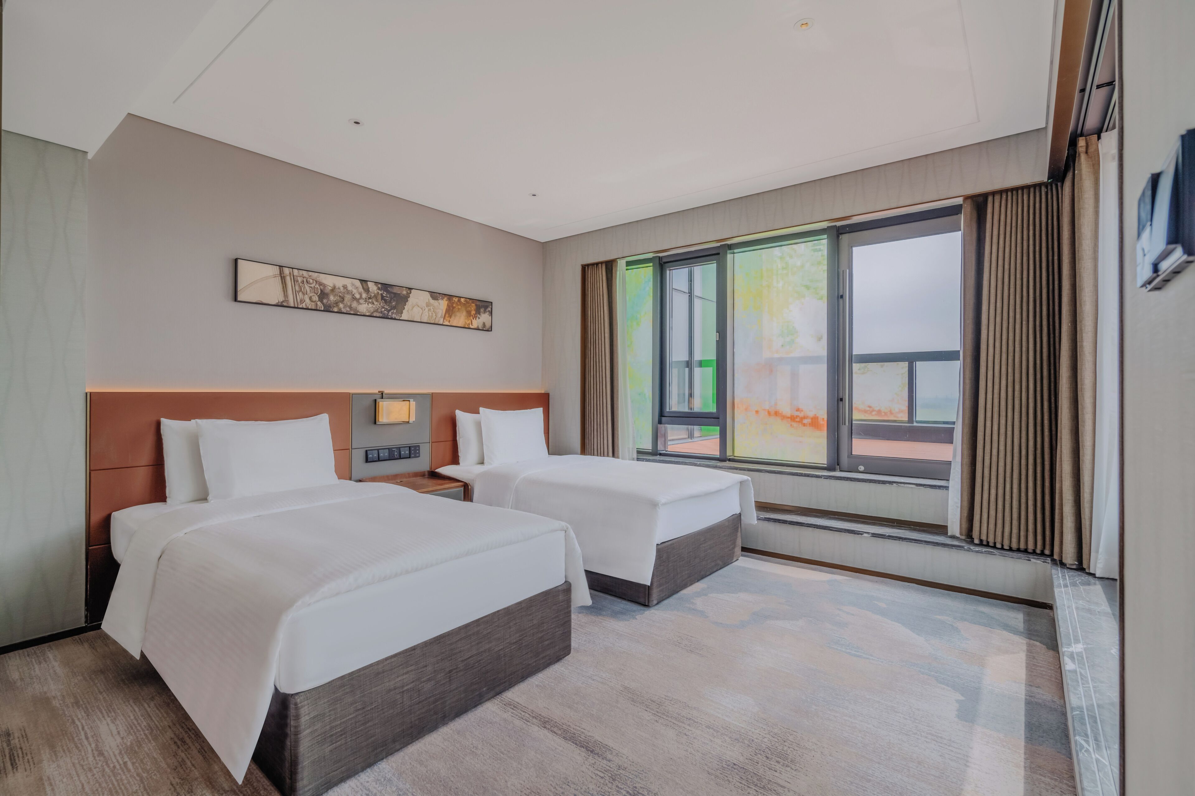 Habitación de hotel con dos camas, un ventanal con cortinas, una obra de arte colgada en la pared y una mesita de noche con lámpara.