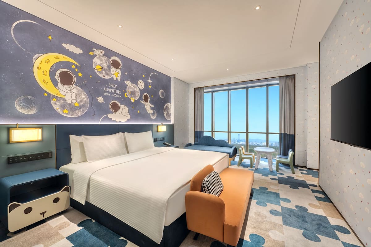 Habitación de hotel con una cama grande, un sofá, una mesita y vistas de la ciudad a través de amplias ventanas.