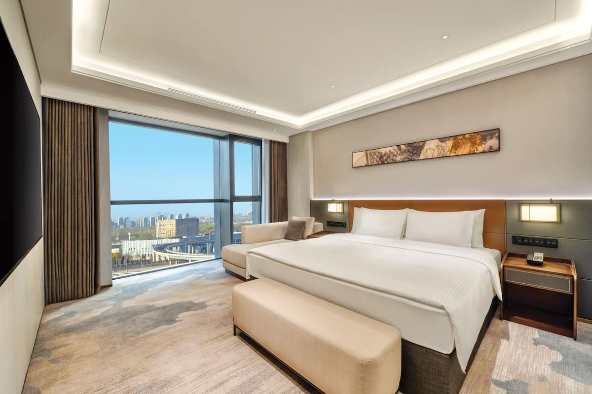 Habitación de hotel moderna con una cama grande, un banco, una mesita de noche y vistas a la ciudad.