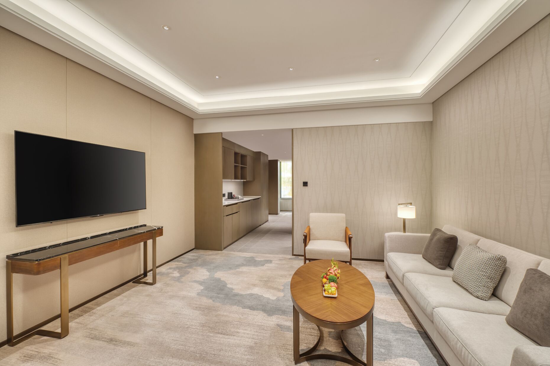 A modern hotel room with a flat-screen TV, a wooden coffee table with fruit, a sofa, and a dining area with a kitchen in the background.
