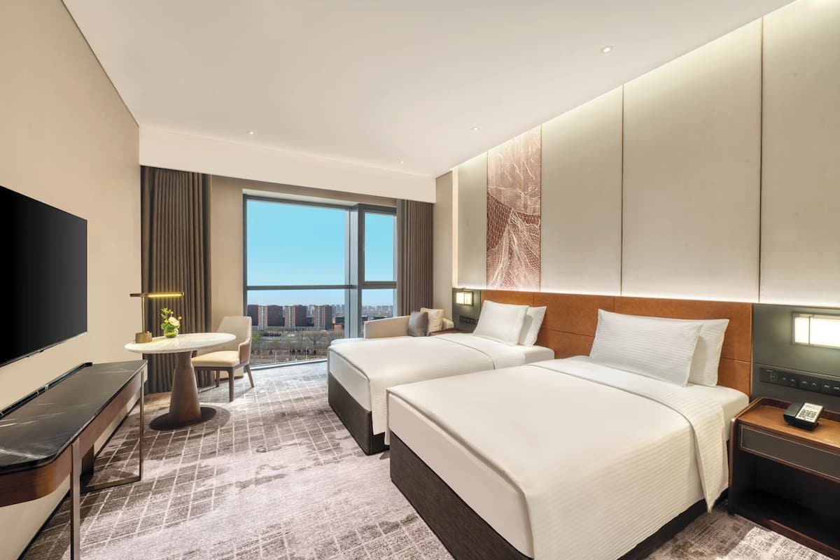 Habitación de hotel con dos camas, un escritorio, televisor y un amplio ventanal con vistas a la ciudad.