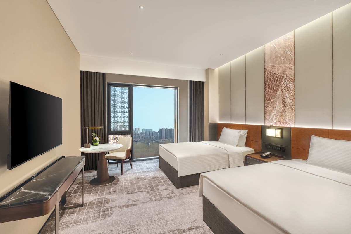 Habitación de hotel con televisor grande, dos camas, escritorio y vista a la ciudad.