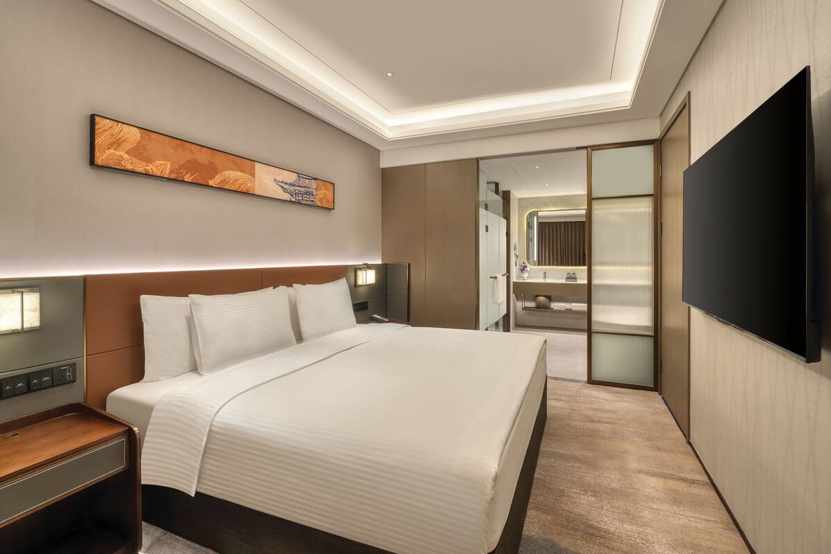 Habitación de hotel con una cama grande, televisión de pantalla plana, mesita de noche y una pintura en la pared, junto con una puerta corredera que da a un baño.