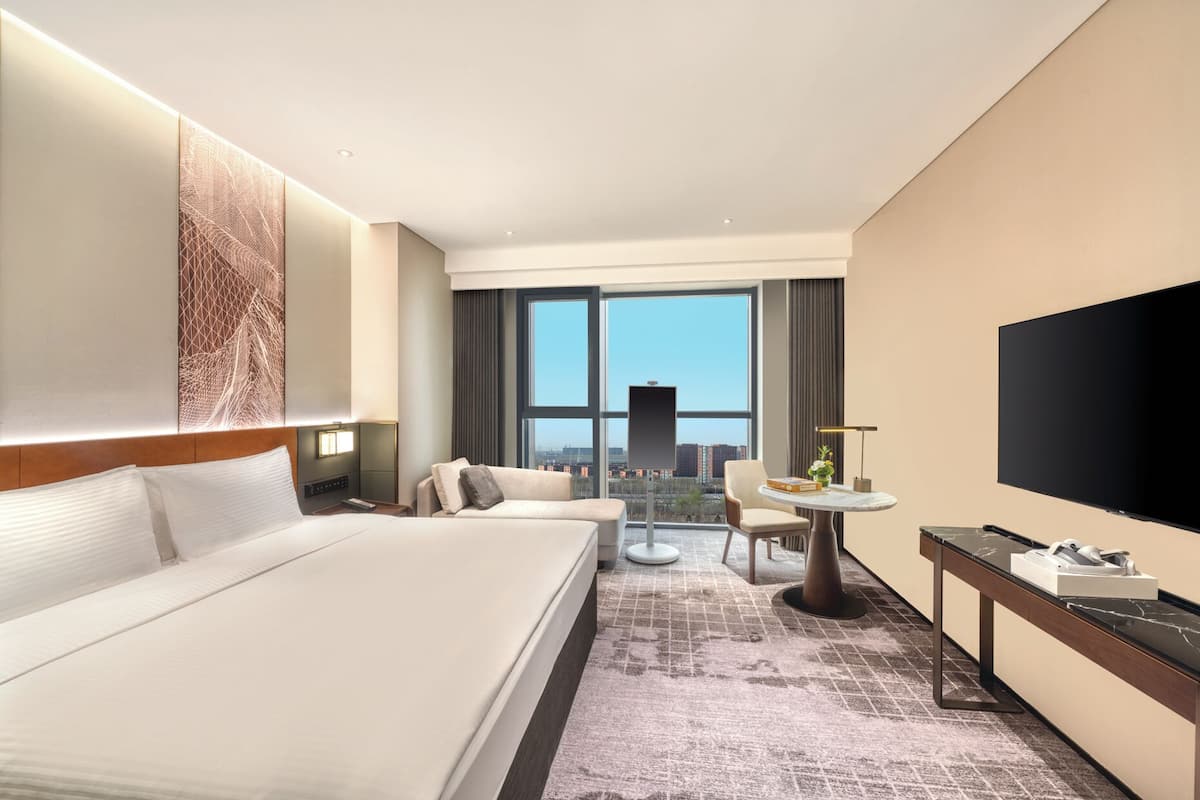 Habitación de hotel con una cama grande, un escritorio con televisor de pantalla plana, una mesa redonda pequeña con un jarrón y vistas de la ciudad a través de amplias ventanas.