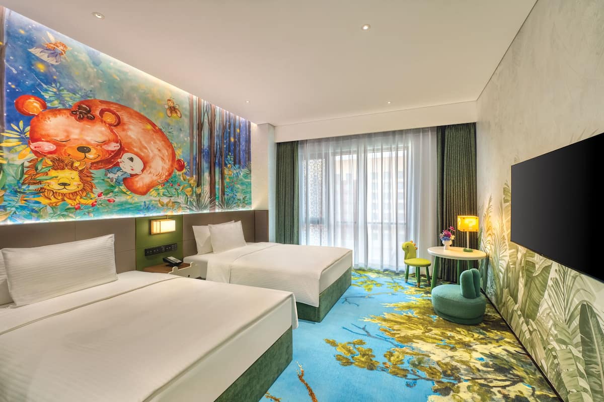 Habitación de hotel con dos camas, un mural grande, un televisor, una mesita con lámpara y una silla verde.