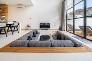 A modern living room with a large sectional sofa, a dining area with a table and chairs, a flat-screen TV, and large windows. - Nunggalan 1 Bedroom Loft Villa with Pool, Walk to Beach (Pecatu)