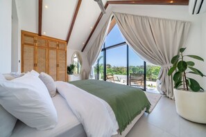 Ein Schlafzimmer mit Bett, großem Fenster, Blick ins Grüne und einer Zimmerpflanze.
