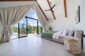 Ein Schlafzimmer mit einem Bett, einem großen Fenster, einem Deckenventilator und Blick auf Grün.