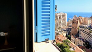 A view through a window with blue shutters, overlooking a coastal cityscape with buildings and the sea. - Appartement 10B - MyResidenced'Azur (Beausoleil)
