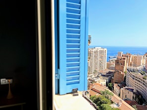 Blick durch ein Fenster mit blauen Fensterläden auf eine Küstenstadt mit Gebäuden und dem Meer.