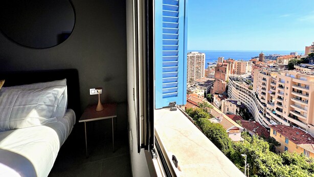 A hotel room with a bed, a small table, a nightstand, and a window with a view of a cityscape and the sea. - Appartement 10B - MyResidenced'Azur (Beausoleil)