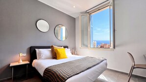 A modern bedroom with a bed, bedside tables, a chair, and a large window offering a city view. - Appartement 10B - MyResidenced'Azur (Beausoleil)