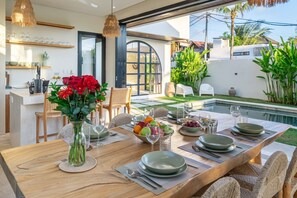 A dining area with a table set for four, surrounded by chairs, with a view of a swimming pool and greenery. - Large 3 Bedroom Villa in Prime Uluwatu with Pool and Free Airport Pickup (South Kuta)