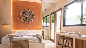 Une chambre d’hôtel moderne avec un lit, un fauteuil en osier, un coin bar et une grande fenêtre.