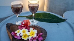 Une table avec deux verres contenant un liquide sombre, un bol en bois rempli de fleurs et le tronc d’un arbre dans un pot.
