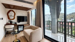 A hotel room with a sofa, a small table, a TV, and a balcony with a view of buildings and trees.