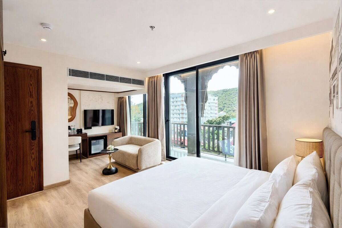 A hotel room with a large bed, a desk with a chair, a TV, and a balcony with curtains.