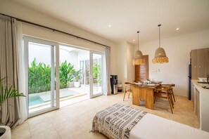 A modern hotel room with a large sliding glass door leading to a balcony, a wooden dining table, and a kitchenette. - 2BR Nunggalan Villa with Pool & Rooftop (South Kuta)