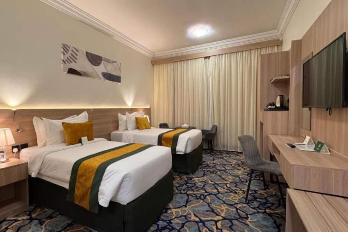 Ein Hotelzimmer mit zwei Betten, einem Schreibtisch, einem Sessel, einem Fernseher und einem gemusterten Teppich.