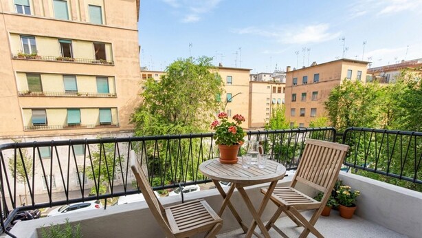 Ein Balkon mit zwei Holzstühlen, einem runden Tisch, Topfpflanzen und Blick auf umliegende Gebäude.