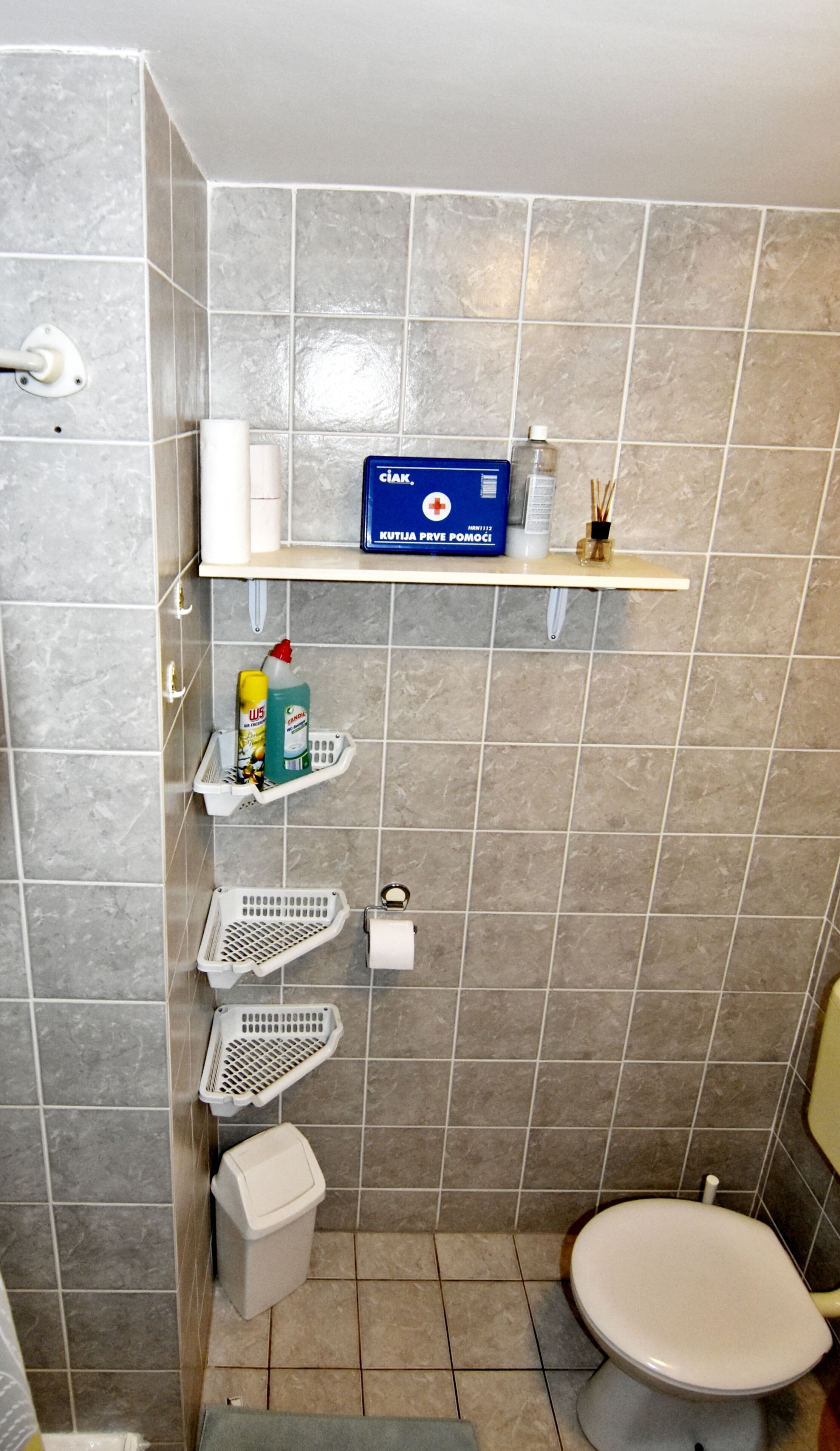 A bathroom with grey tiles, a toilet, a shelf with toiletries, and a trash bin.