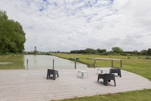 A wooden deck with outdoor seating, a small table, and a boat on a calm water body. - Holiday Home 'Le T'chot Pierre 1' with Lake View, Shared Garden and Wi-Fi (Saint-Quentin-en-Tourmont)
