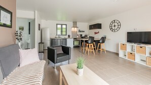 Un salon moderne avec un canapé, un coin repas avec des chaises, une cuisine équipée de meubles et un téléviseur posé sur un meuble.