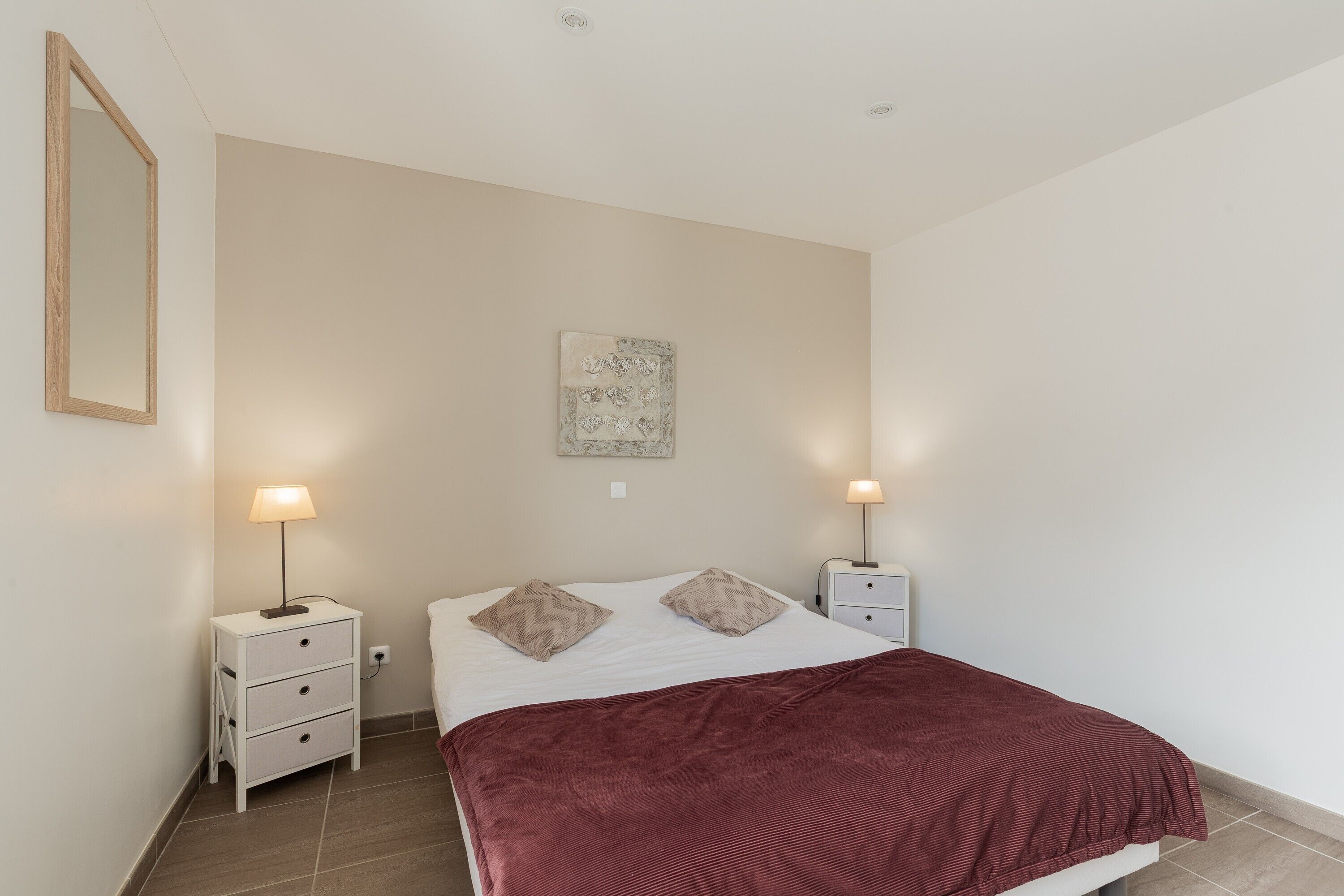 Een slaapkamer met een bed, twee nachtkastjes, twee lampen, een kunstwerk aan de muur en een rode spreidekker.