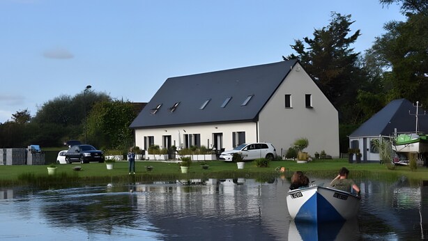 Une maison avec un toit en pente, un étang devant et un bateau avec des personnes à bord.