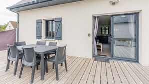 Un espace extérieur couvert aménagé pour les repas, avec une table et des chaises, une porte-fenêtre coulissante donnant sur une cuisine et une terrasse en bois.