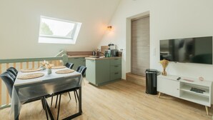 A dining area with a table, chairs, a kitchen, a TV, and a trash can. - Vacation Rental Le Filou (Saint-Quentin-en-Tourmont)