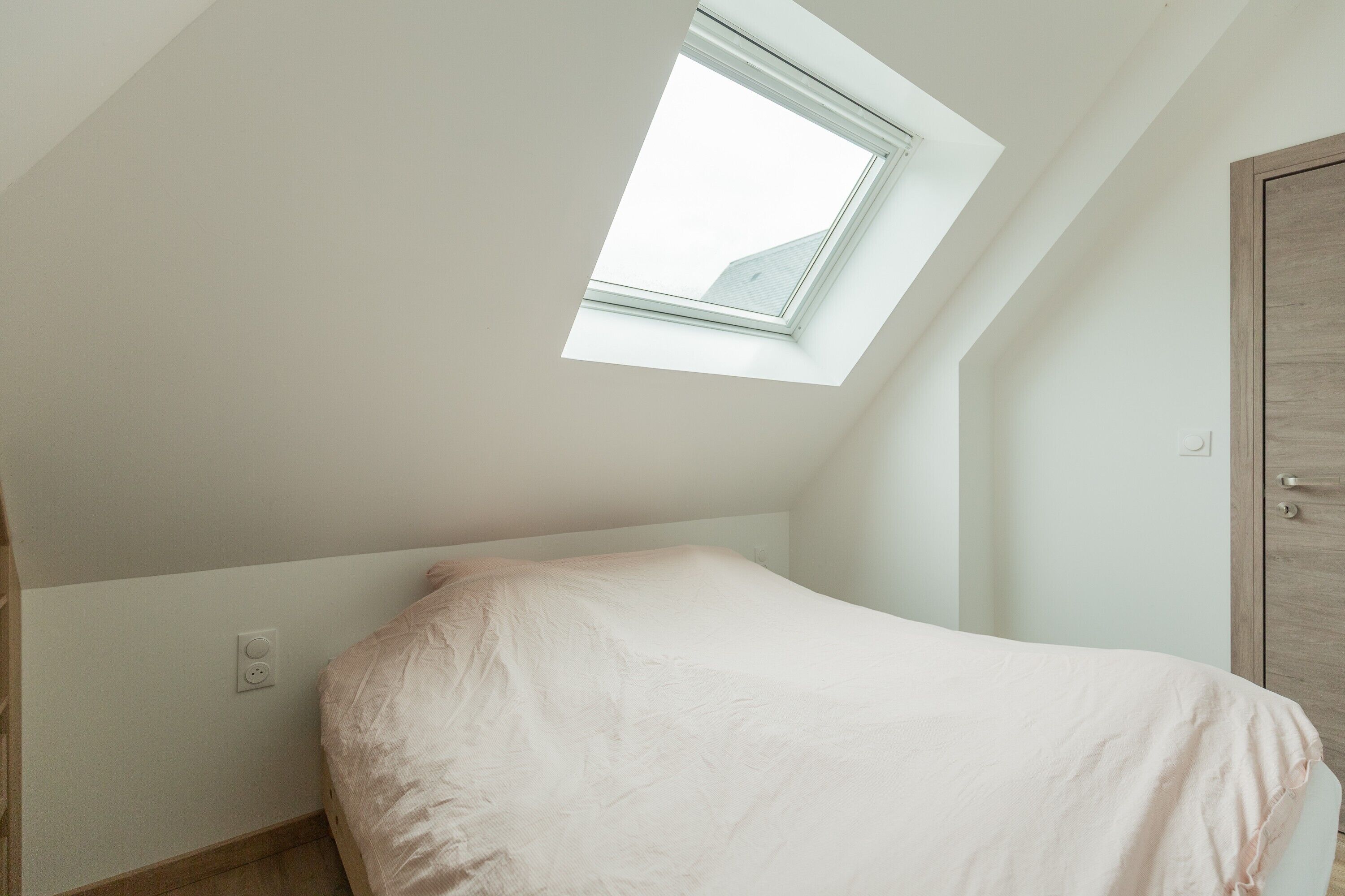 A room with a sloped ceiling, a skylight, a bed, a wooden wardrobe, and a wall socket.
