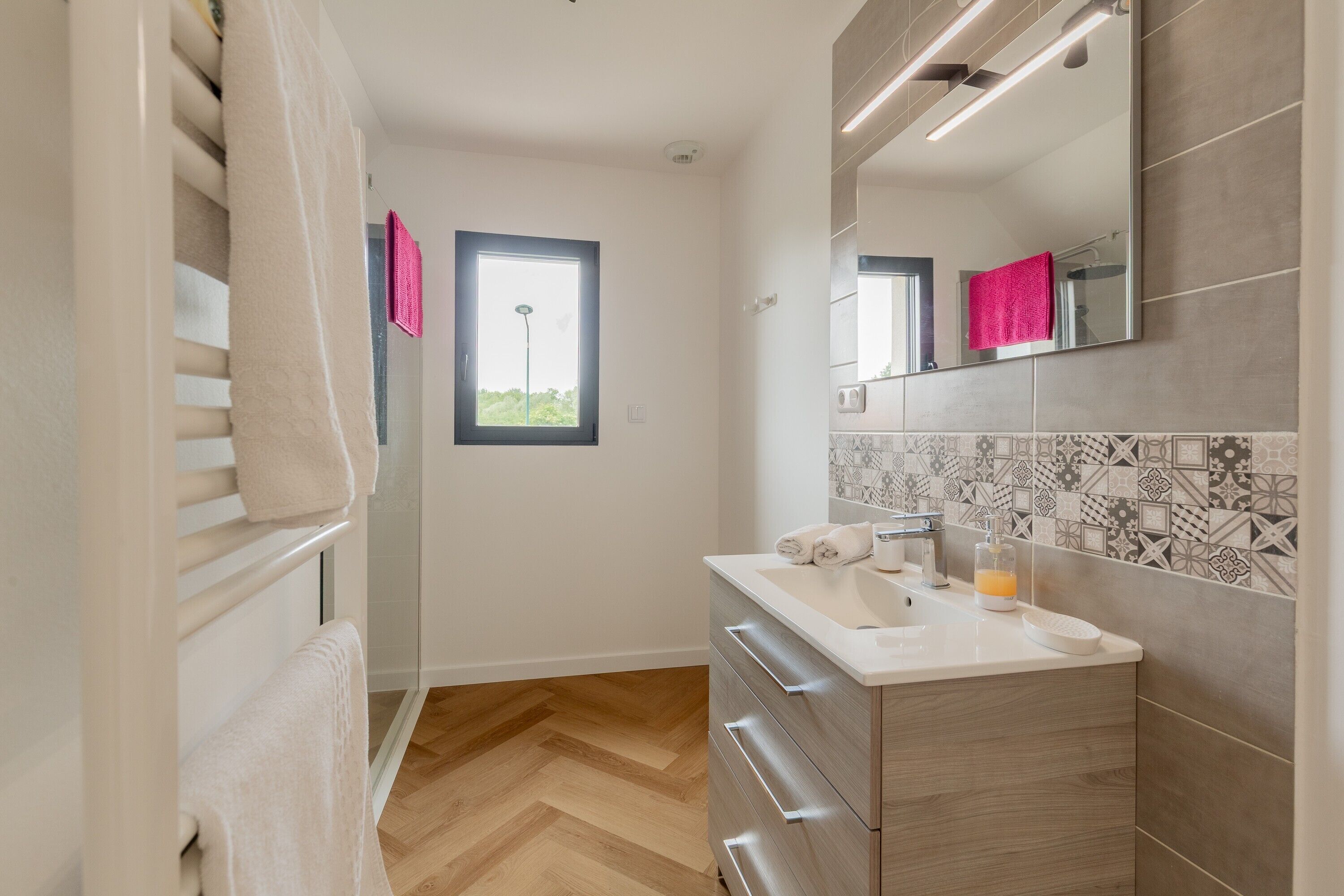 Une salle de bain moderne avec un plancher en bois, un lavabo blanc, un miroir et une fenêtre.