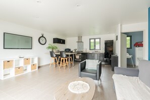 A modern living room with a sofa, a dining area with chairs, a kitchen with cabinets, and a TV mounted on the wall. - Holiday Home 'Le T'chot Pierre 2' with Lake View, Private Terrace and Wi-Fi (Saint-Quentin-en-Tourmont)