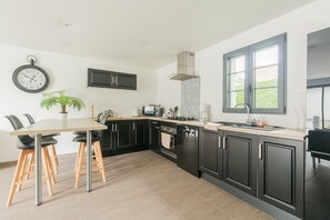 A modern kitchen with dark cabinetry, a dining table with chairs, a clock, and a window. - Holiday Home 'Le T'chot Pierre 2' with Lake View, Private Terrace and Wi-Fi (Saint-Quentin-en-Tourmont)