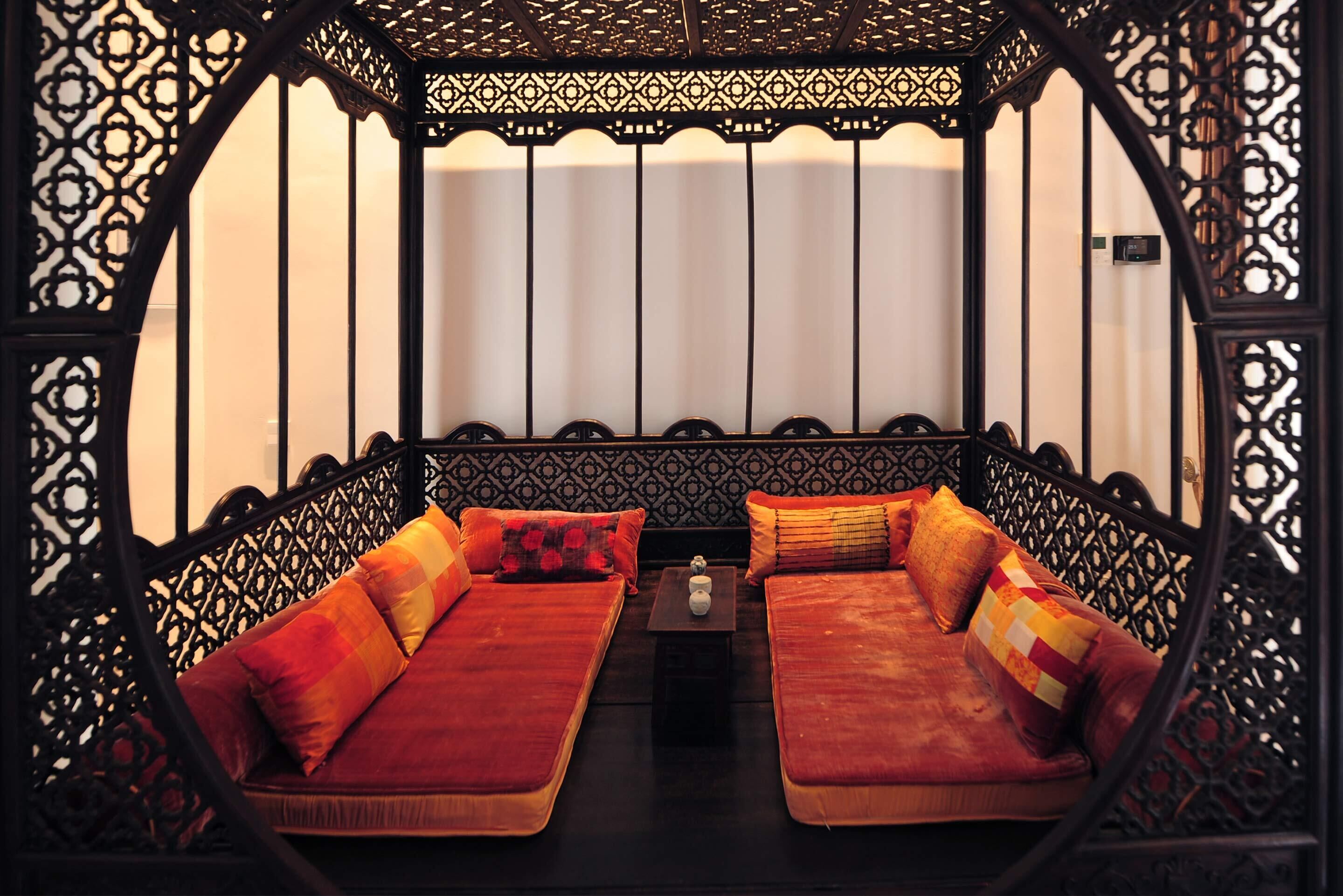 A room with a low, ornate wooden bench with cushions, a small table with a vase, and a patterned wall.