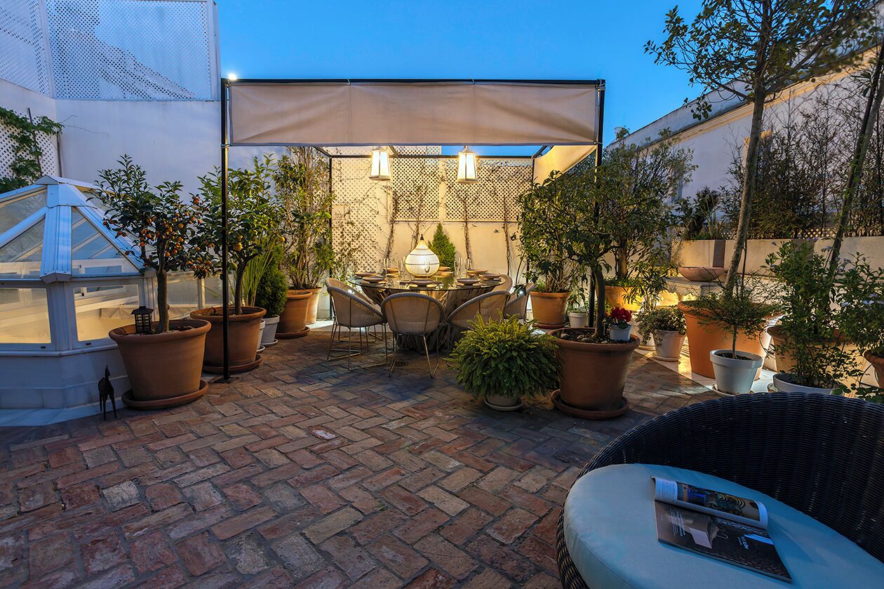 Een terras met een tafel en stoelen, omringd door potplanten en een pergola met verlichting.