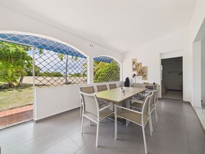 Un espace repas moderne avec une grande table et des chaises, une vue sur un jardin à travers une fenêtre grillagée et une porte donnant sur une autre pièce.