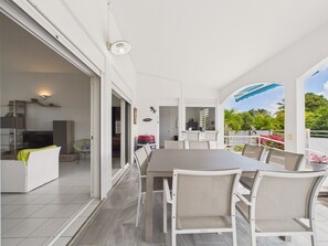 A modern outdoor dining area with a large table and chairs, a kitchen in the background, and a view of a white fence and greenery. - Charming house with pool near golf course 8 guests (Saint-François)