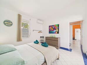 A bedroom with a bed, a dresser, a window with blinds, a colorful painting, and a door leading to another room. - Charming house with pool near golf course 8 guests (Saint-François)