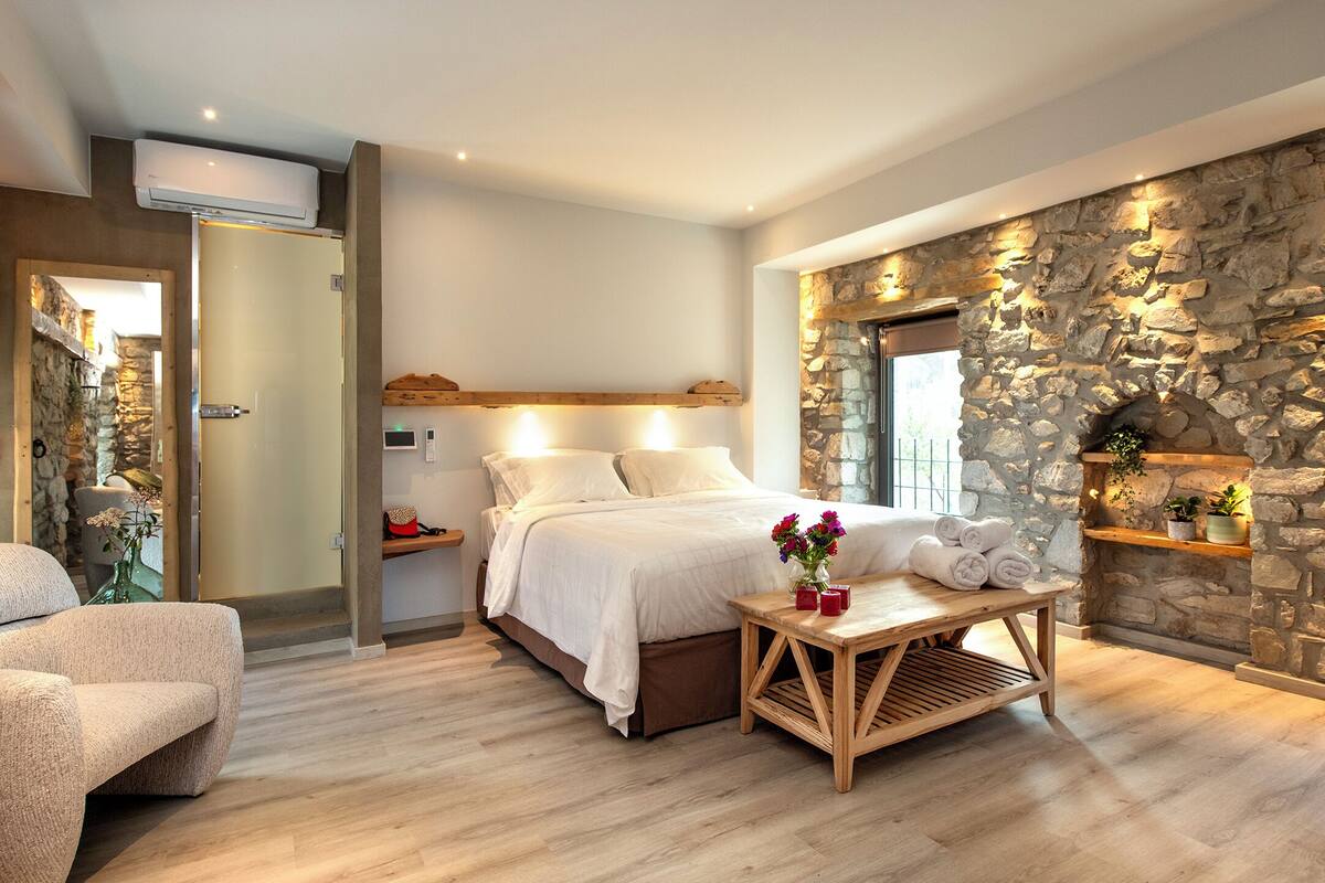 Un dormitorio con pared de piedra, una cama con ropa blanca, una mesita de noche de madera y una pequeña mesa con toallas y flores.