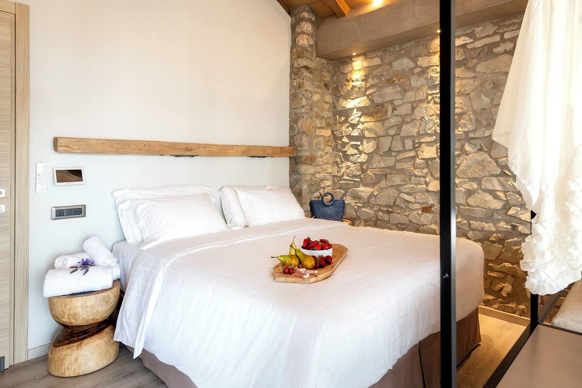 Una cama con ropa blanca, una tabla de frutas y quesos, una pared de piedra y un cabecero de madera.