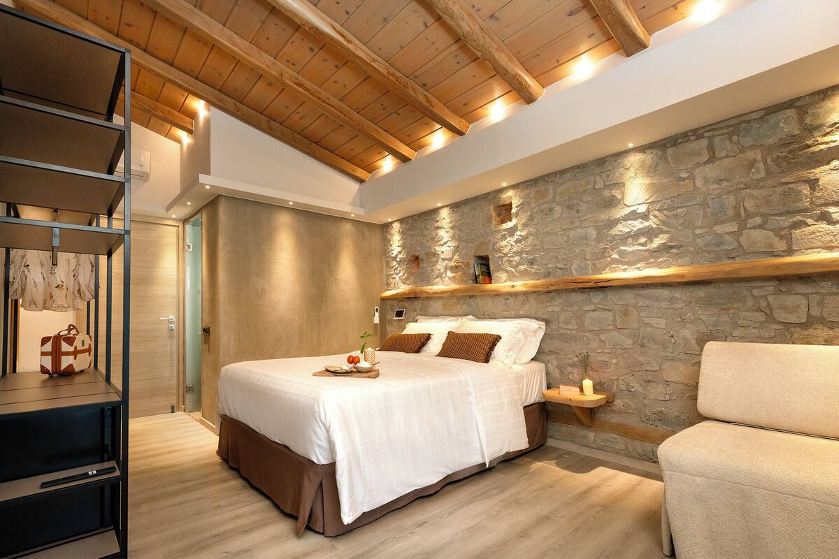 Un dormitorio con pared de piedra, vigas de madera en el techo, una cama con ropa de cama blanca, una mesita de noche y una silla.